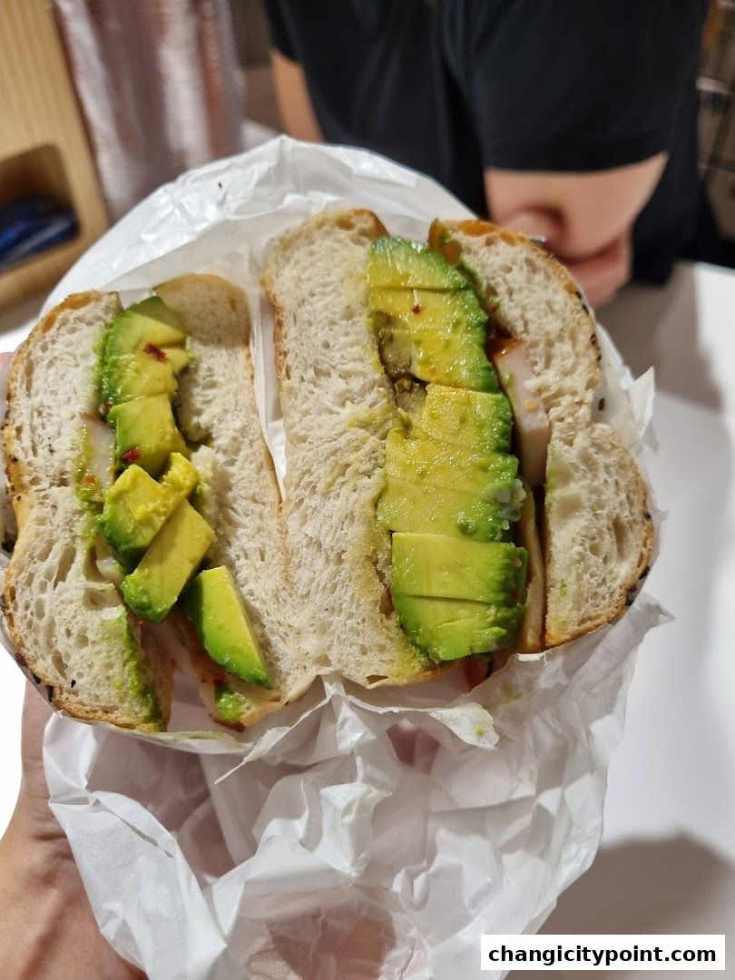 A hand holds a delicious avocado sandwich on a rustic bread, wrapped in white paper.