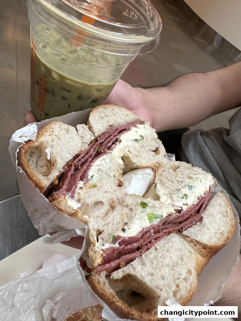 A person holds a large bagel sandwich filled with meat and cream cheese, next to a green iced drink.