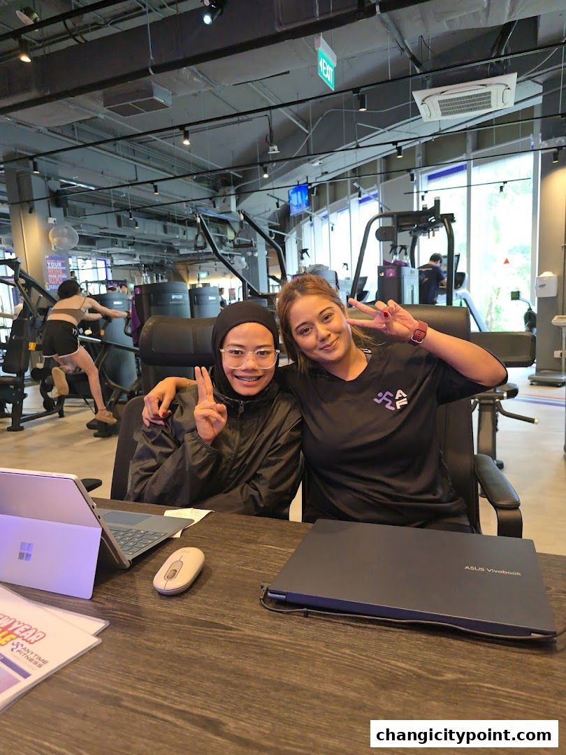 Two smiling women posing at a desk in a brightly lit gym with exercise equipment.