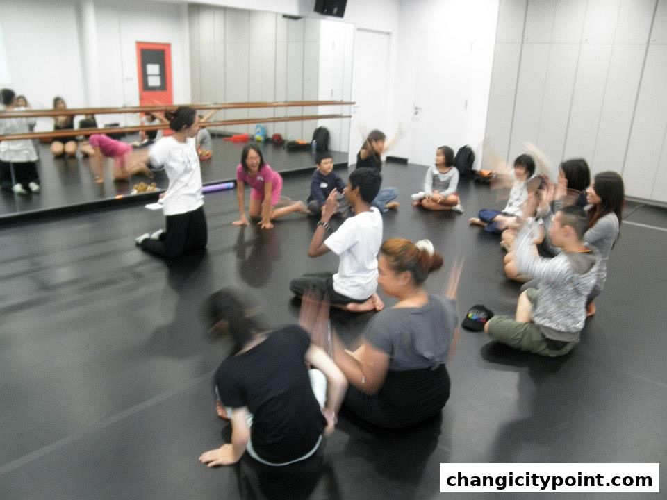 A group of people, likely students, are sitting in a circle on a dance studio floor.