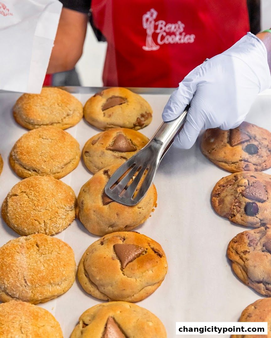 A gloved hand uses tongs to pick up a freshly baked cookie from a tray.