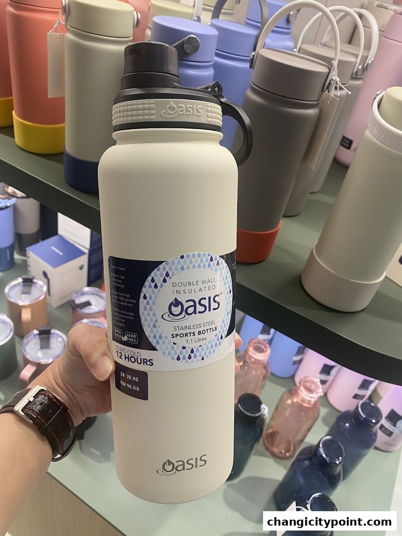 A hand holds a cream-colored Oasis stainless steel sports bottle, with other bottles displayed behind.