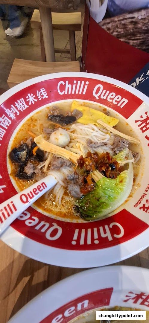 A bowl of rice noodles with meat, vegetables, and a spoon from Chilli Queen.