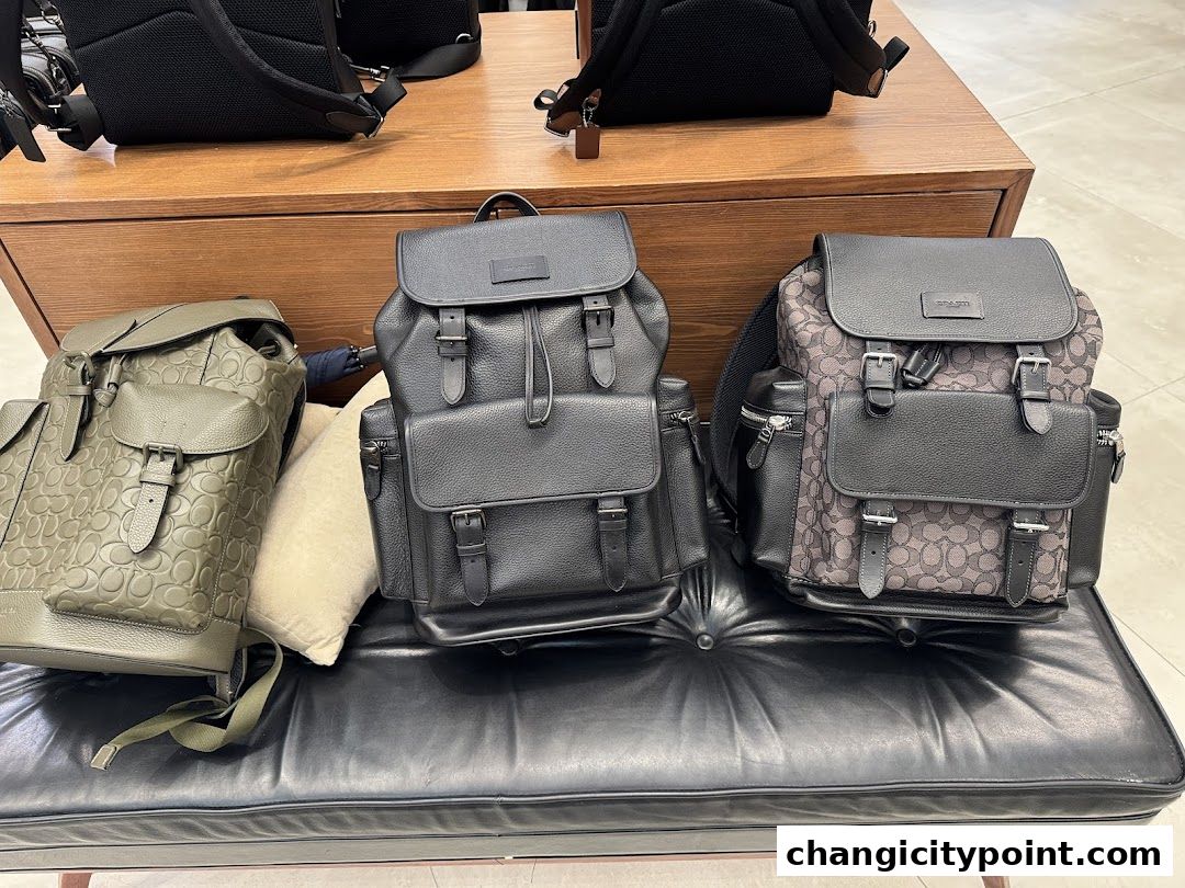 Three Coach backpacks displayed on a black leather bench.