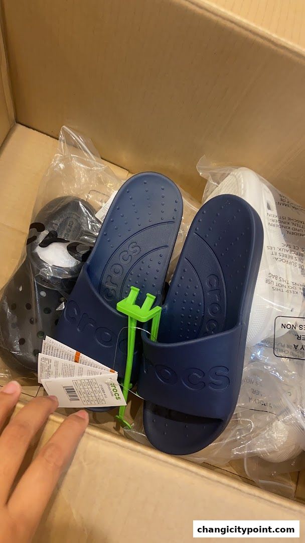 A box filled with various Crocs sandals, including navy blue slides and black clogs.