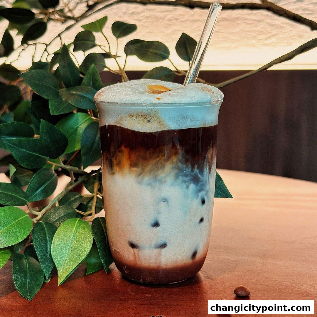 A layered iced coffee drink with foam and a straw, next to green leaves.