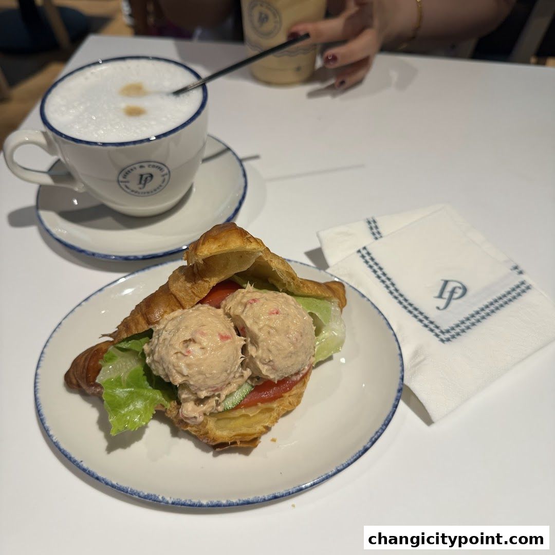 A croissant sandwich with tuna salad and a latte on a white table.