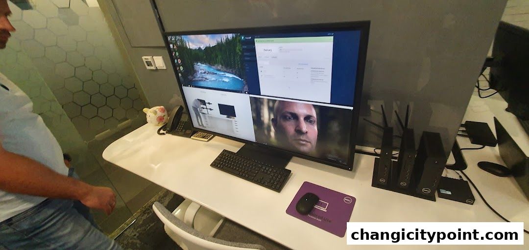 A person stands near a desk with a large monitor displaying various content, including a landscape and a face.