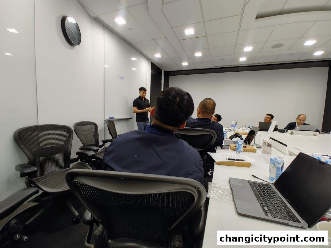 A presenter stands in front of a whiteboard while attendees sit at a conference table.