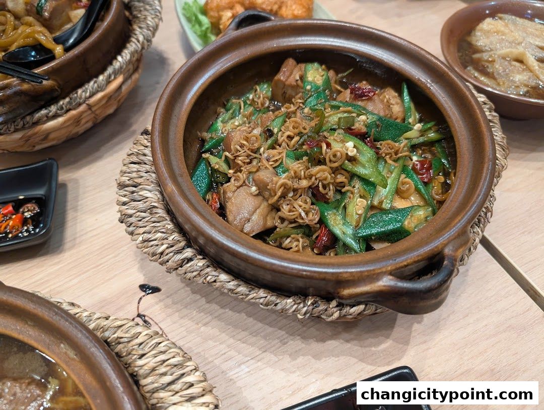 A close-up of a clay pot dish with okra, meat, and noodles, served on a table.