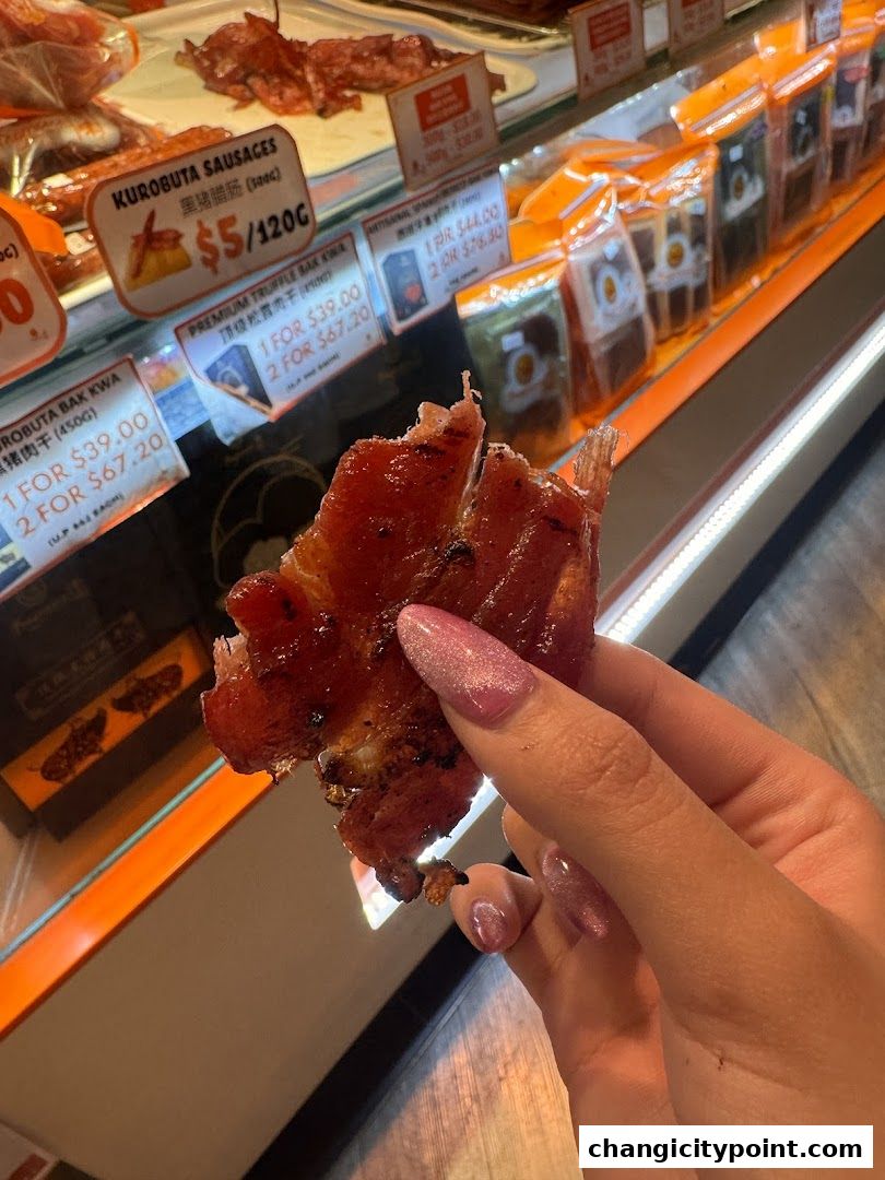 A hand holds a piece of bak kwa in front of a display of meat products.