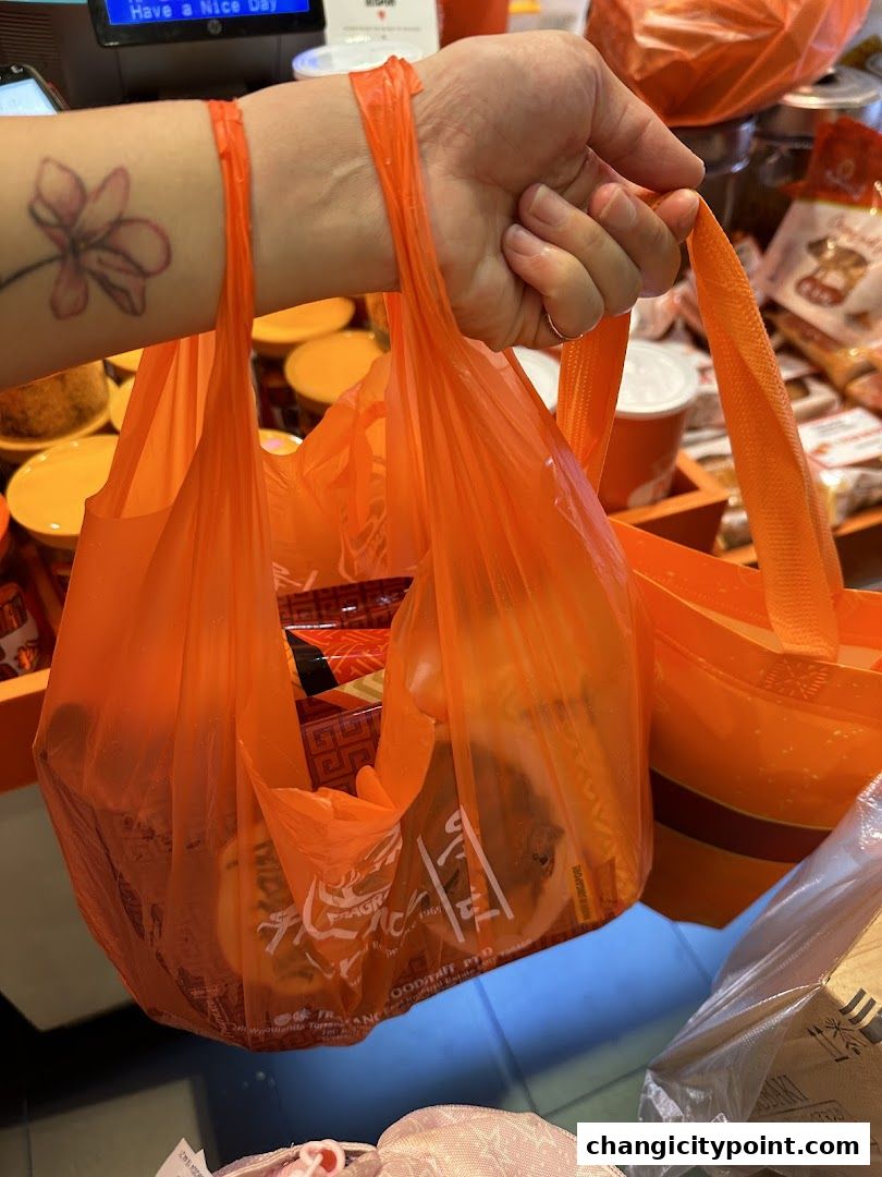 A hand holds two orange plastic bags filled with food items from Fragrance Bak Kwa.