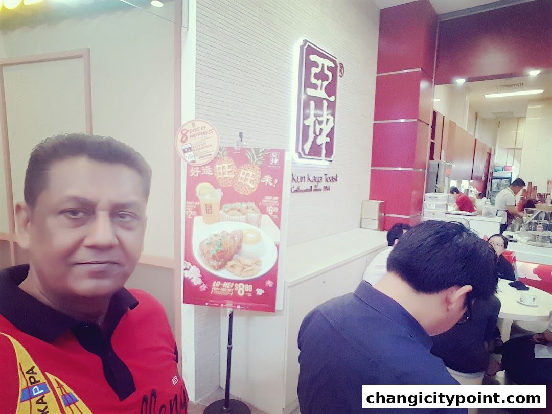 A man in a red shirt poses in front of a restaurant with a promotional poster.