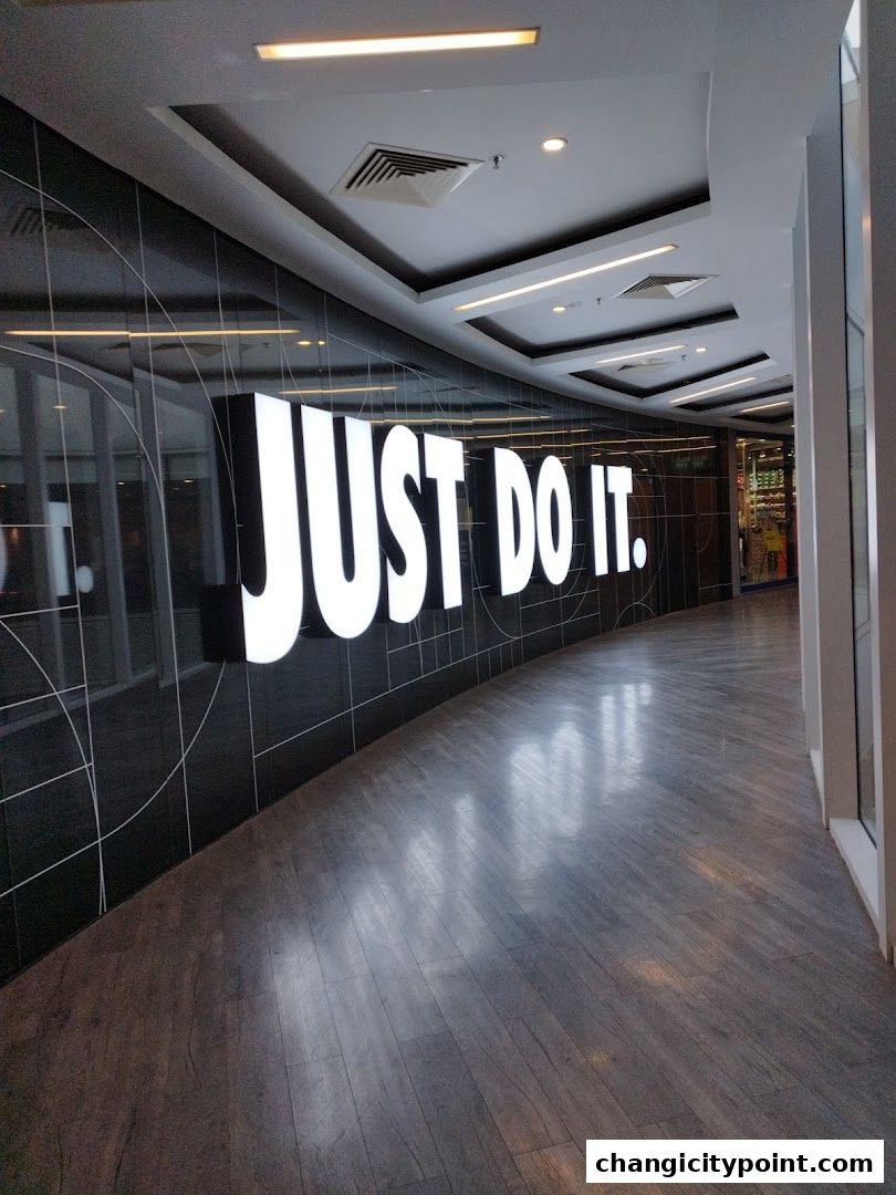 A large illuminated 'JUST DO IT.' sign on a black tiled wall in a modern mall hallway.