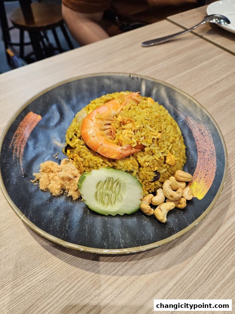 A plate of yellow fried rice with a shrimp, cucumber slice, cashews, and shredded meat.