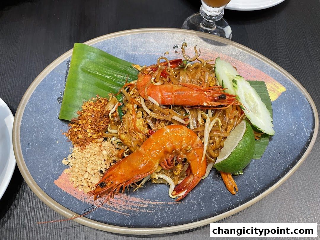 A plate of Pad Thai with large prawns, lime, cucumber, and crushed peanuts.