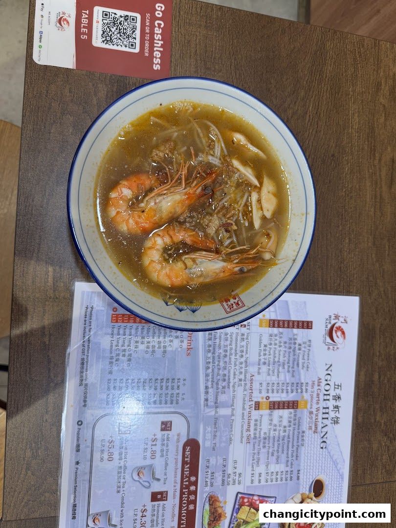 A bowl of prawn noodles sits next to a menu with various food and drink options.