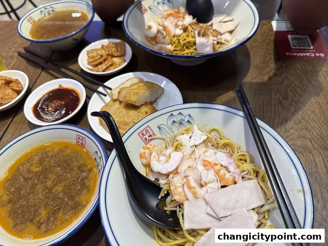Bowls of prawn noodles with various side dishes and dipping sauces on a wooden table.