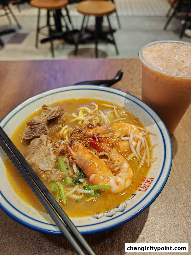 A bowl of prawn noodles with a drink on a wooden table.
