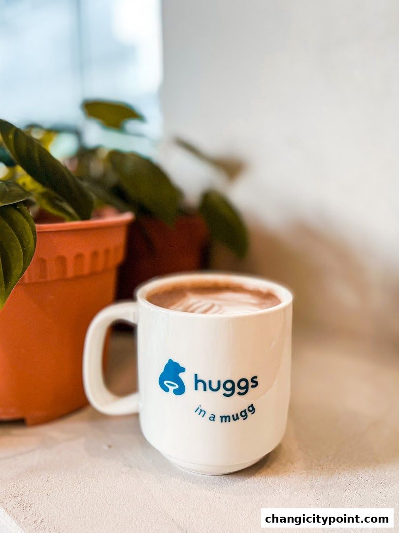 A white mug with the Huggs Coffee logo and a latte art design sits next to potted plants.