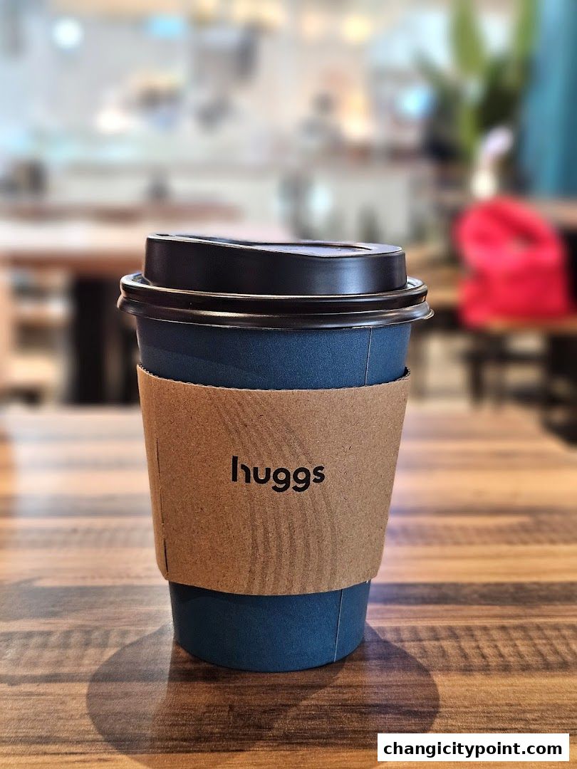 A blue Huggs Coffee cup with a brown sleeve sits on a wooden table.