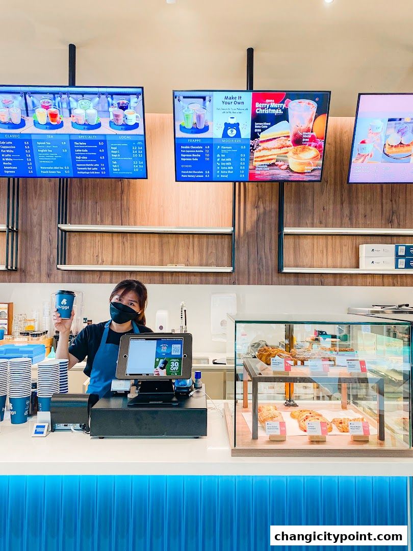 A barista holds a Huggs Coffee cup at the counter with digital menus and pastries displayed.