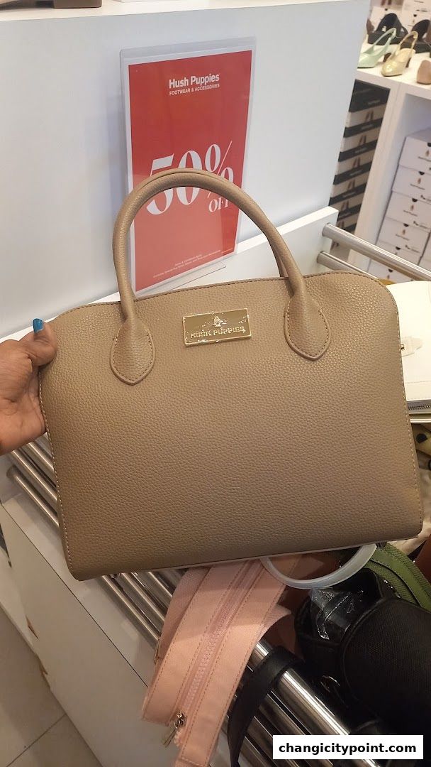 A person holds a tan Hush Puppies handbag in front of a 50% off sale sign.