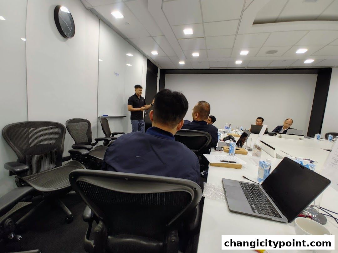 A presenter stands in front of a whiteboard in a meeting room with attendees seated.