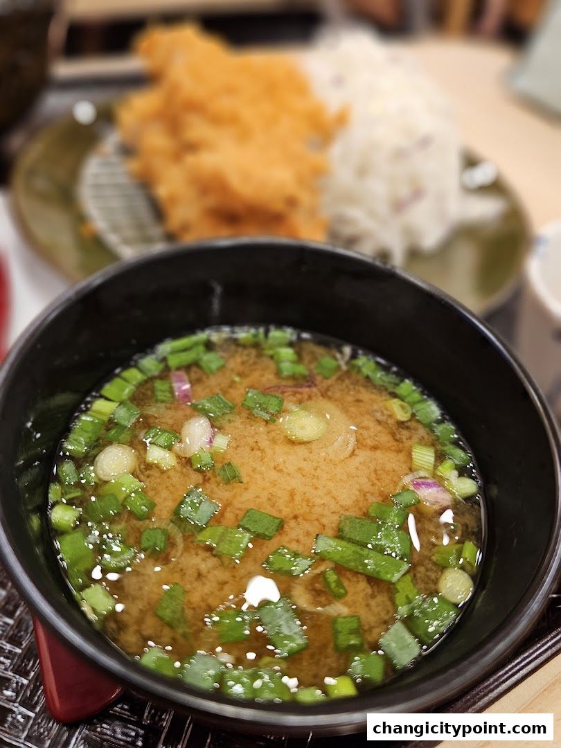 A close-up of miso soup with chopped green onions and a blurred katsu and rice dish.