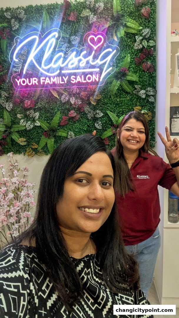 Two smiling women in a salon with a neon sign and floral wall.