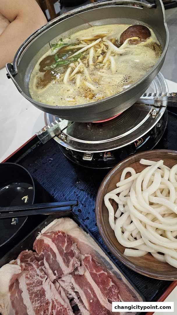 A hot pot meal with thinly sliced meat, udon noodles, and broth with vegetables.