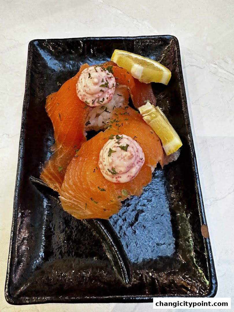 Smoked salmon sushi with creamy topping and lemon slices on a black plate.