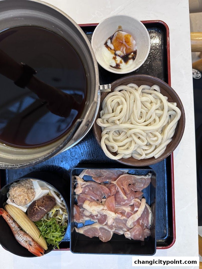 A close-up of a hot pot meal with udon noodles, raw meat, vegetables, and a soft-boiled egg.