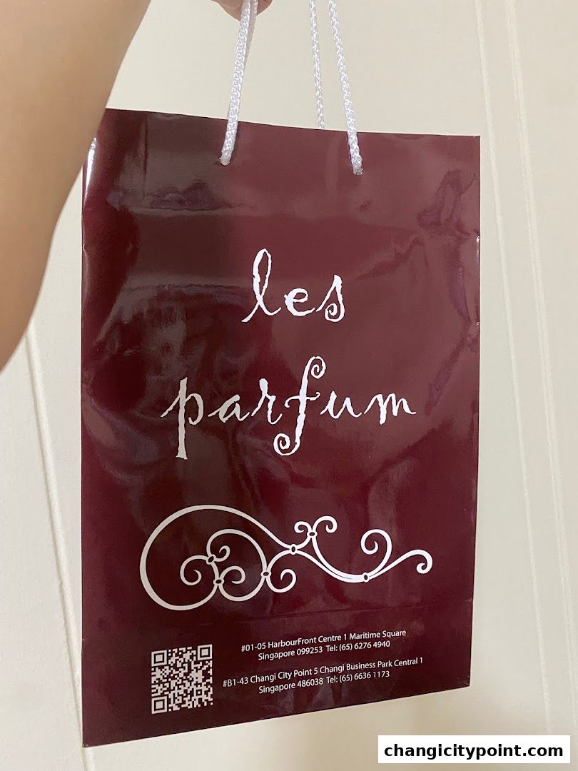 A person holds a burgundy shopping bag with 'les parfum' logo and store details.