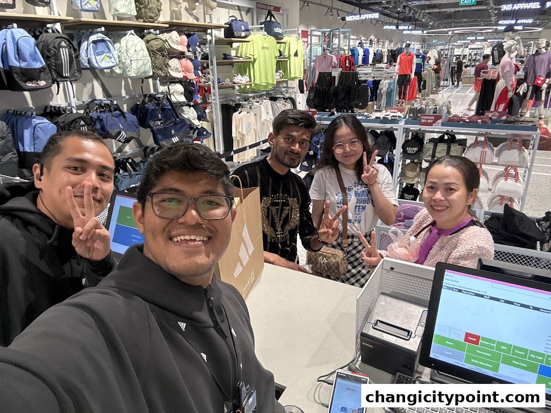 A group of smiling staff members pose for a selfie in a well-stocked Adidas store.