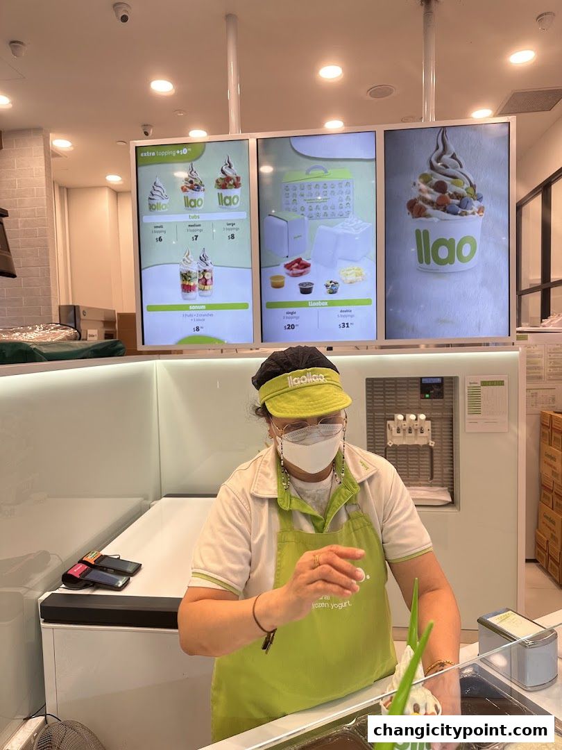 A frozen yogurt shop employee stands behind the counter with digital menus displaying frozen yogurt options.