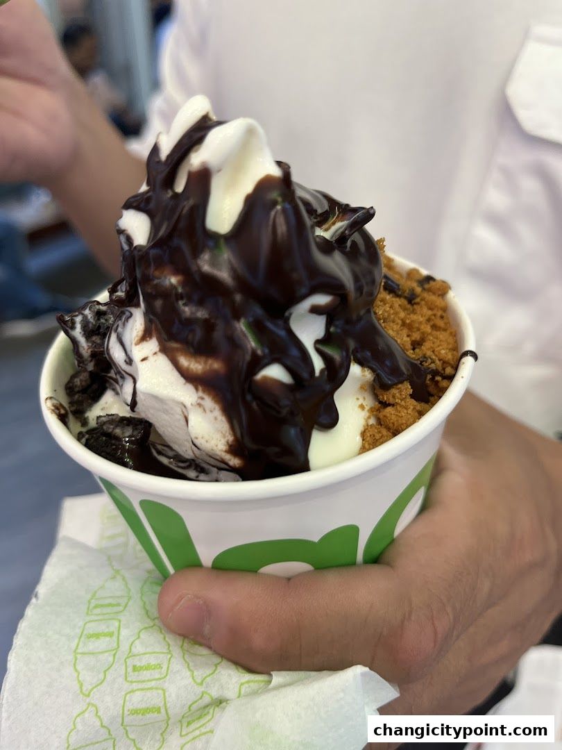 A hand holds a cup of frozen yogurt topped with chocolate sauce and cookie crumbles.