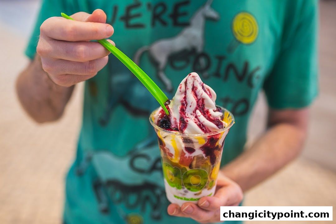 A person holds a cup of frozen yogurt with fruit and sauce, being eaten with a green spoon.