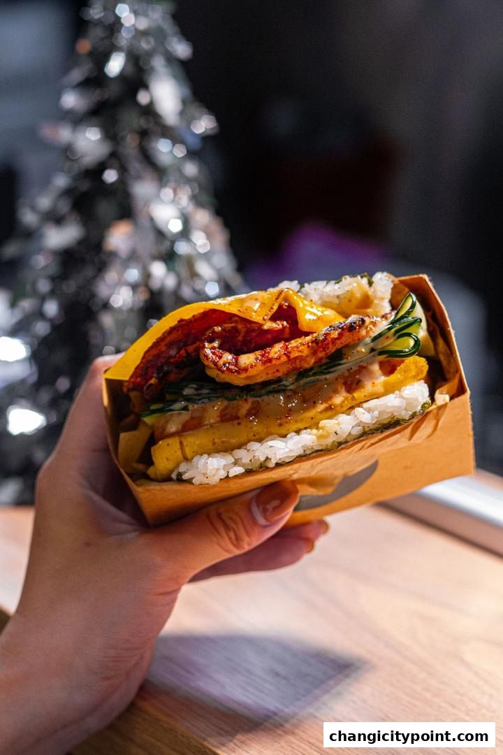 A hand holds a delicious onigiri sandwich with rice, egg, cucumber, and grilled meat.