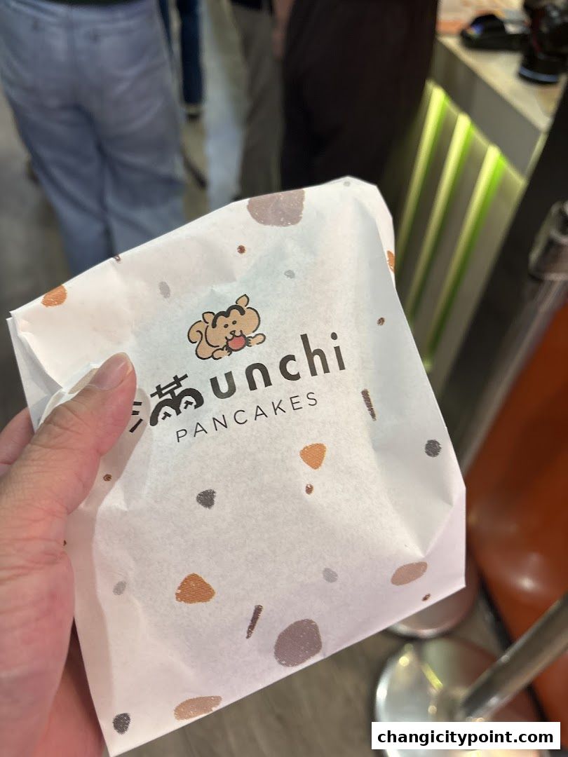 A hand holds a paper bag with the Munchi Pancakes logo and a cute cartoon character.