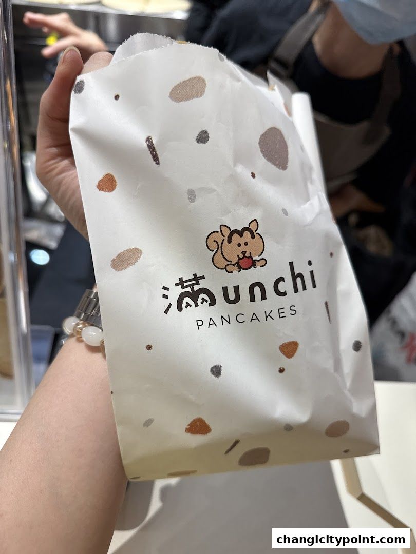A person holds a branded paper bag from Munchi Pancakes with a cute squirrel logo.