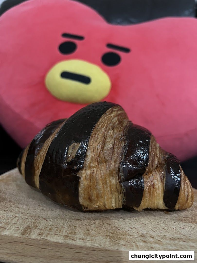 A chocolate croissant sits on a wooden board with a large, pink character plush in the background.