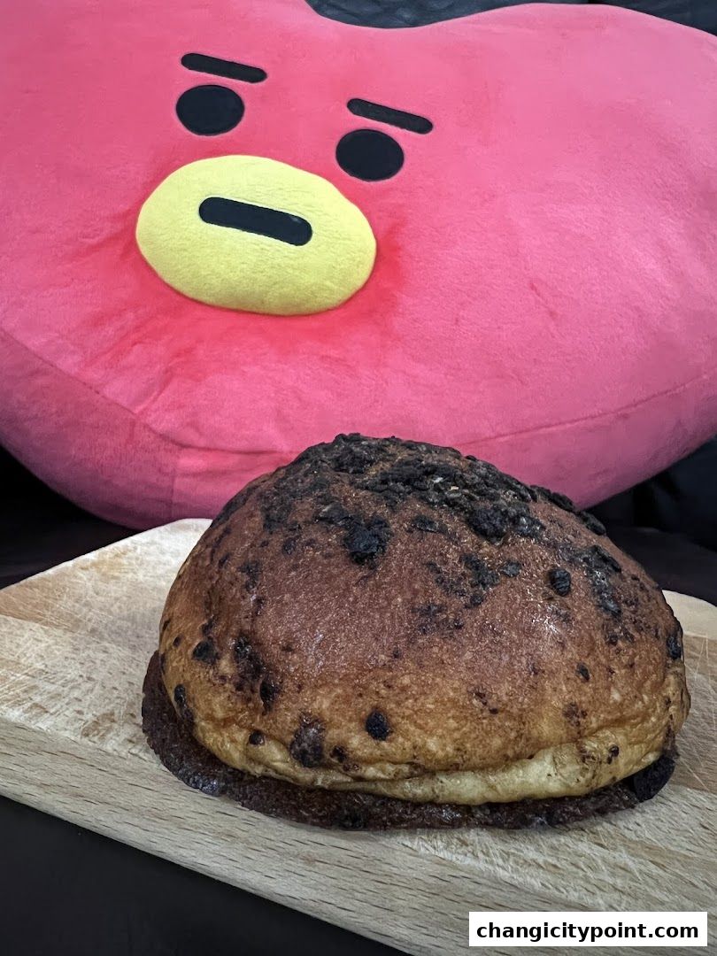 A freshly baked bun with dark toppings sits on a wooden board, with a large pink character cushion behind it.
