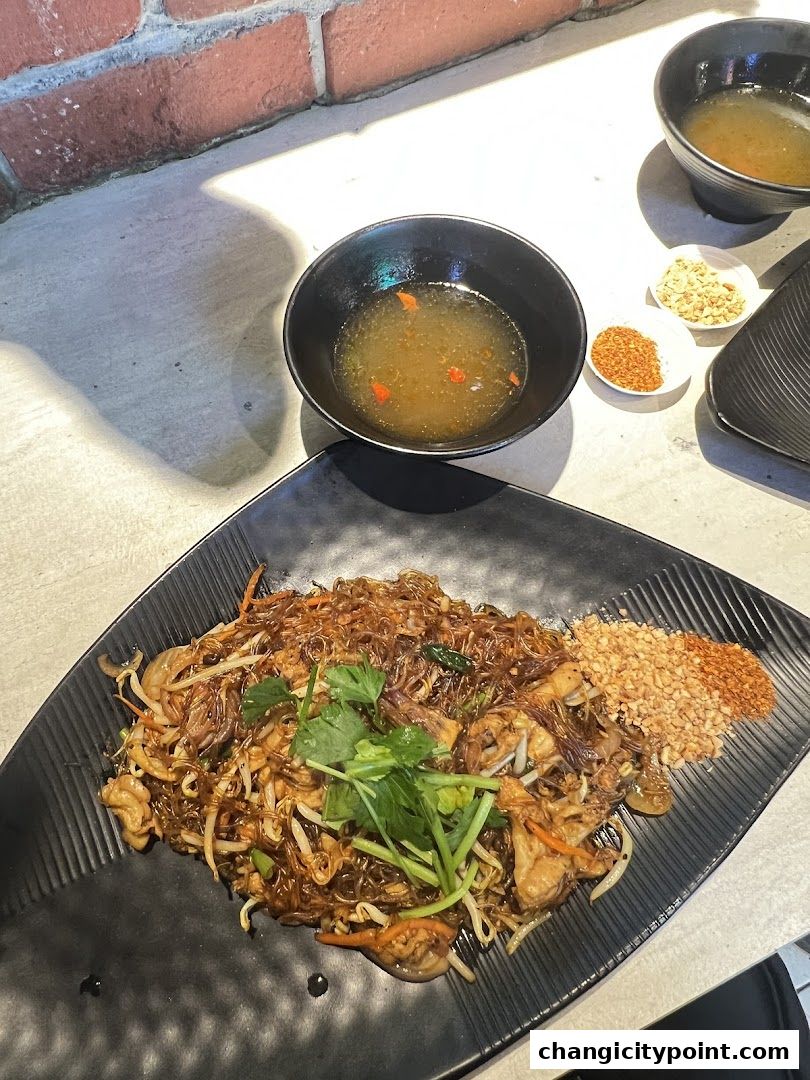A plate of Pad Thai noodles with a side of chili flakes and crushed peanuts.