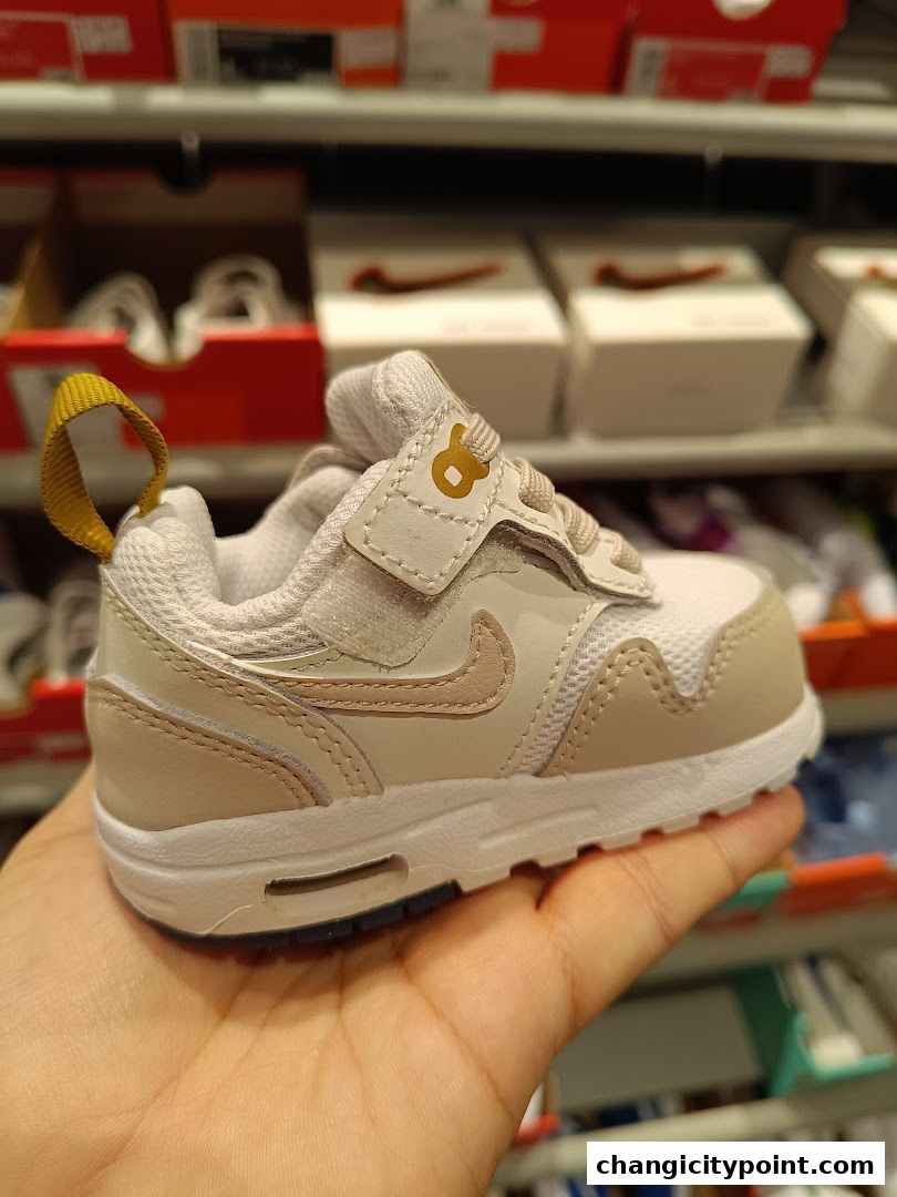 A hand holds a small, light-colored Nike sneaker with a gold accent.