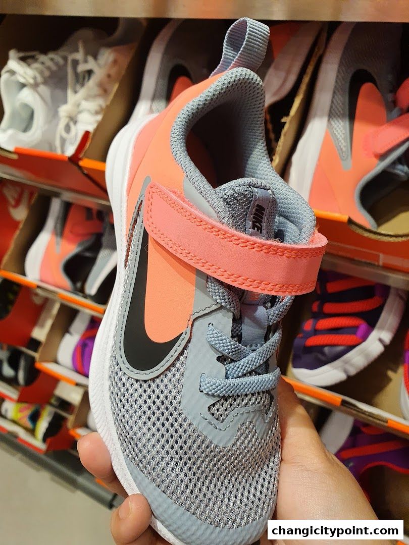 A hand holds a pink and grey Nike sneaker with other shoes displayed on shelves.