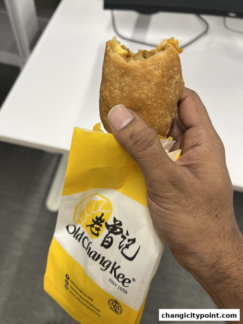 A hand holds a partially eaten Old Chang Kee curry puff in its branded yellow packaging.