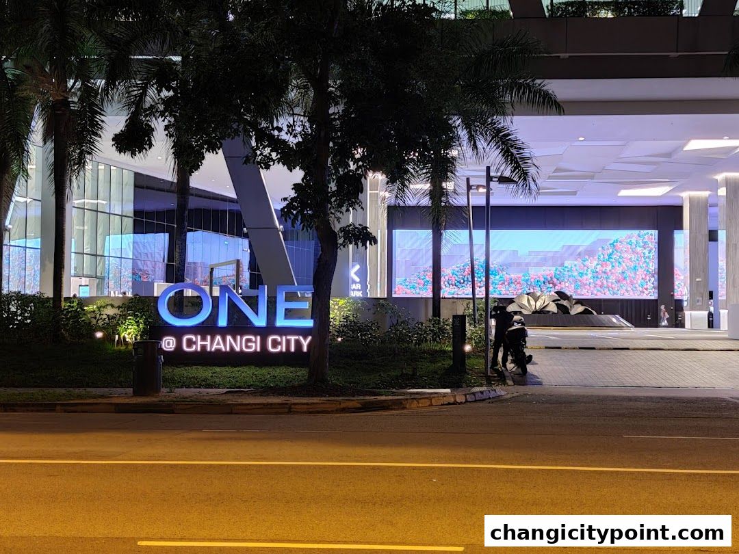 The illuminated sign for ONE @ Changi City stands before a modern building entrance at night.