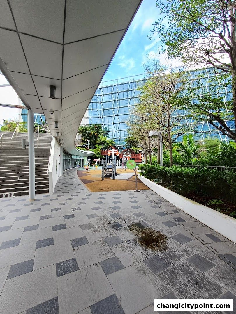 A modern walkway leads to a playground with a glass building in the background.