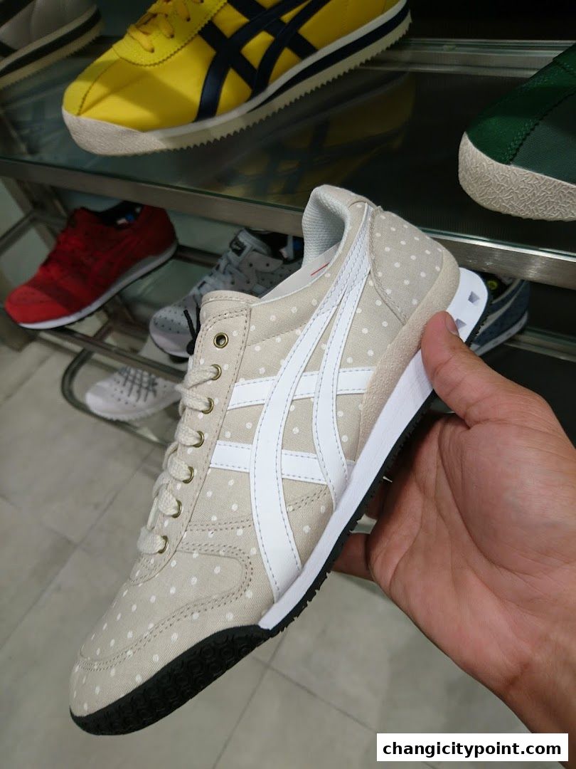 A hand holds a beige polka dot Onitsuka Tiger sneaker with other shoes on display.